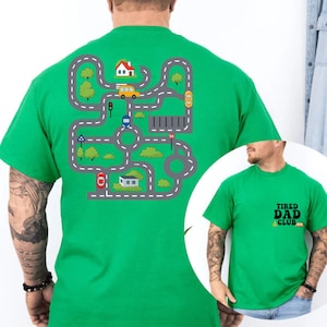 May include: A bright green t-shirt featuring a road map design with cars, houses, and trees. The shirt also has the text "TIRED DAD CLUB" on the front. The design is printed on a green t-shirt.