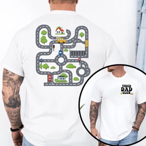May include: White t-shirt featuring a colorful road map design on the back with cars, houses, and trees. The front of the shirt has the text "TIRED DAD CLUB" in black and green.