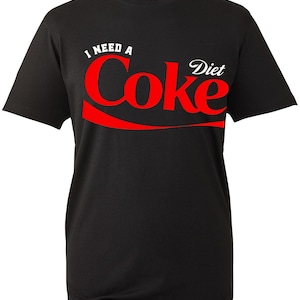 May include: A black short-sleeved t-shirt featuring the white text I NEED A and the word Diet in white, with the word Coke printed in large red letters across the chest, accompanied by a red swoosh graphic underneath the brand name.
