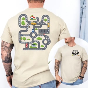May include: Beige t-shirt with a colorful road map design on the back, featuring cars, houses, and trees. The front of the shirt has the text "TIRED DAD CLUB" in black. A fun and playful design for dads.
