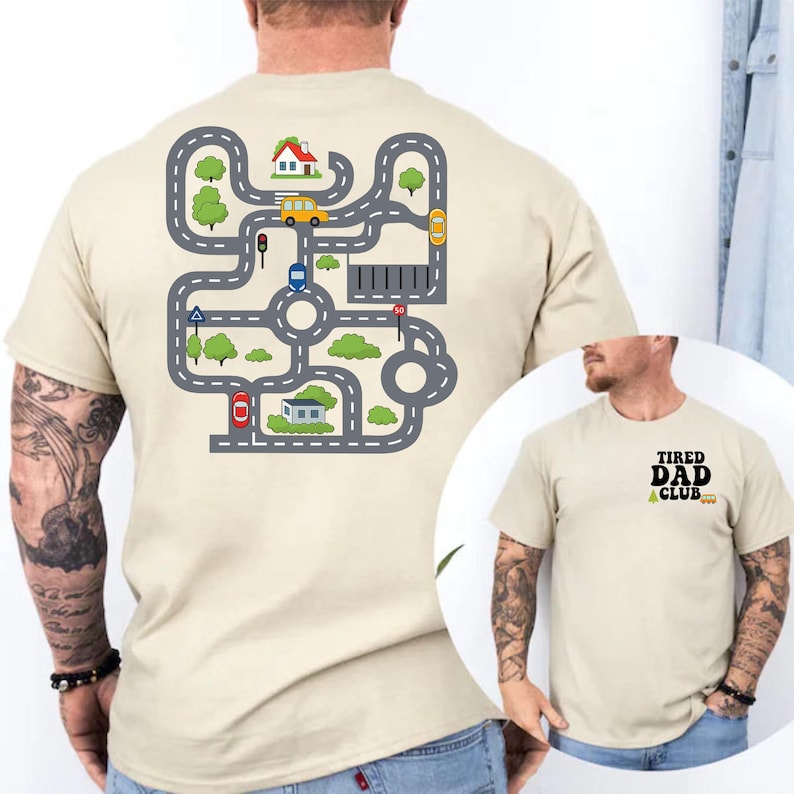 May include: Beige t-shirt with a colorful road map design on the back, featuring cars, houses, and trees. The front of the shirt has the text "TIRED DAD CLUB" in black. A fun and playful design for dads.
