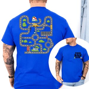 May include: A royal blue t-shirt featuring a road map design with cars, houses, and trees. The shirt also has the text "TIRED DAD CLUB" on the front. The design is printed on the back of the shirt.