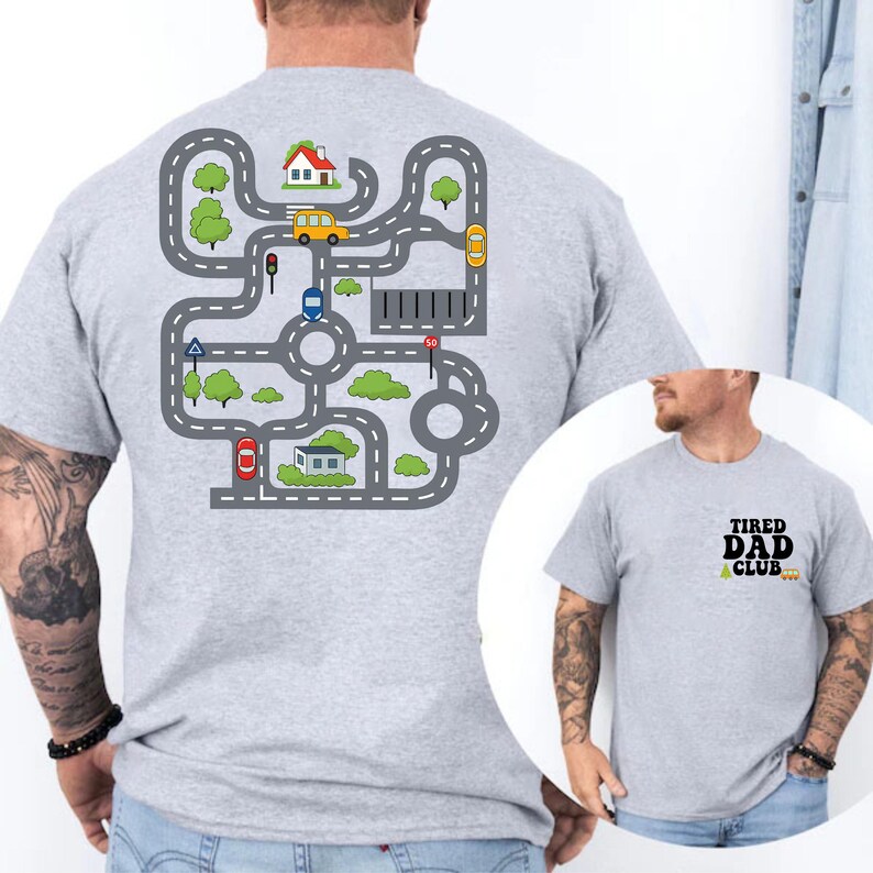 May include: A light gray t-shirt with a road map design on the back, featuring cars, houses, and trees. The front of the shirt has the text "TIRED DAD CLUB" in black and green.