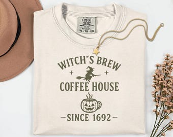Puede incluir: Camiseta color crema con el texto "Witch's Brew Coffee House Since 1692" en verde oscuro. El diseño incluye una bruja en una escoba y una taza de café con forma de calabaza. Un collar con colgante de estrella dorada está sobre la camiseta.