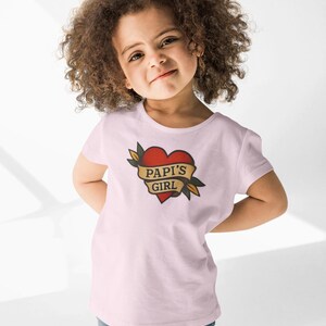 May include: A light pink t-shirt featuring a red heart tattoo design with the words "PAPI'S GIRL" on a banner. The shirt is worn by a young child with curly brown hair and blue jeans.