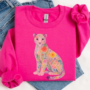 May include: A bright pink sweatshirt featuring a floral leopard design. The leopard is adorned with colorful flowers and leaves. The sweatshirt is paired with blue jeans, creating a casual and stylish look.