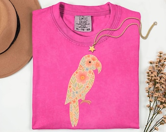 May include: A bright pink t-shirt with a floral parrot design. The parrot is adorned with peach and blue flowers and green leaves. A gold star pendant necklace is draped across the shirt. The shirt has a Comfort Colors tag, size XL-TG-EG.
