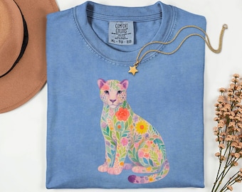 May include: A light blue Comfort Colors t-shirt featuring a floral leopard design. The leopard is adorned with colorful flowers and leaves. A gold star pendant necklace rests on the shirt. The shirt is size XL-TG-EG.