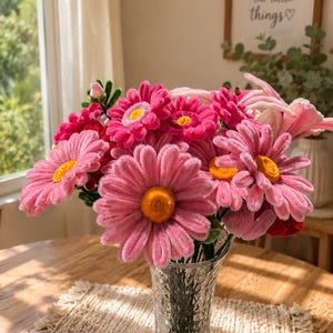 Op de afbeelding: Een boeket van roze en fuchsia bloemen, lijkend op madeliefjes, in een heldere glazen vaas. De bloemen hebben gele centra en staan op een houten tafel met een geweven placemat. Een ingelijste print met de tekst "Enjoy the little things" is zichtbaar.