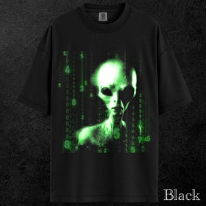 May include: Black t-shirt featuring a green alien graphic with a matrix-style background. The alien has large eyes and a pale green complexion. The word "Black" is printed at the bottom of the shirt.