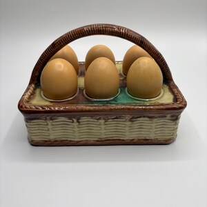 May include: A ceramic egg holder shaped like a basket with a handle. The holder has six slots for eggs and is decorated with brown, green, and white glaze.