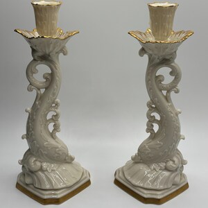 May include: A pair of ivory porcelain candle holders with gold accents. Each holder features a stylized dolphin design with intricate details and a scalloped base.