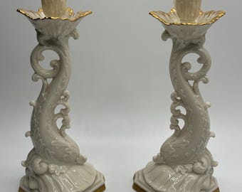 May include: A pair of ivory porcelain candle holders with gold accents. Each holder features a stylized dolphin design with intricate details and a scalloped base.