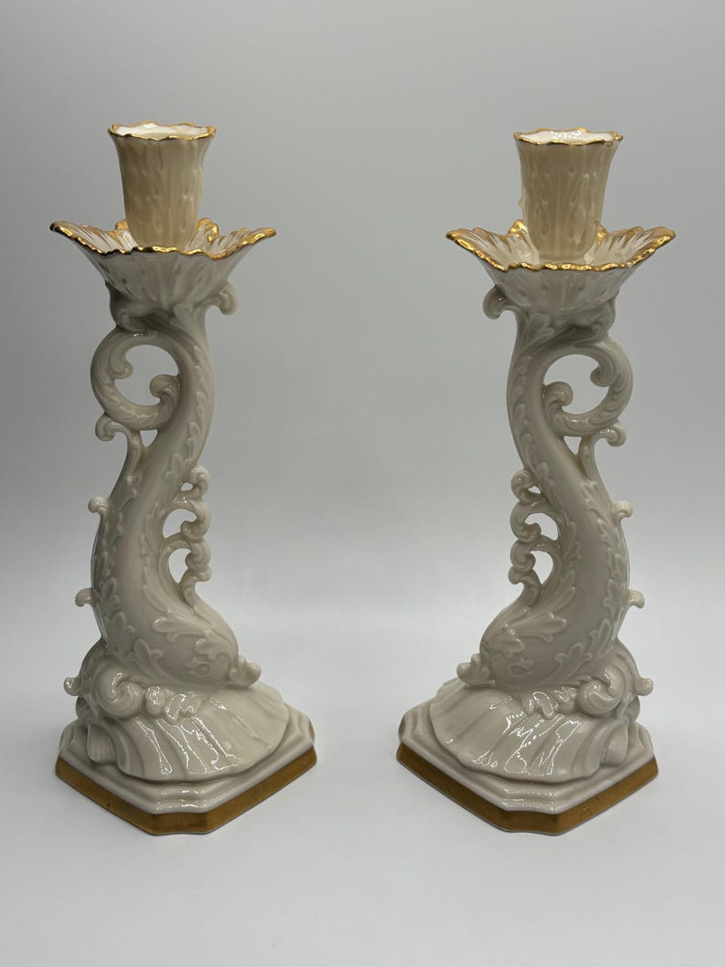 May include: A pair of ivory porcelain candle holders with gold accents. Each holder features a stylized dolphin design with intricate details and a scalloped base.