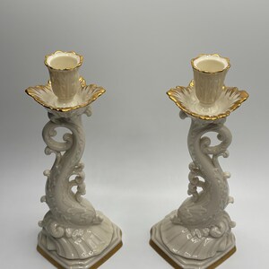 May include: A pair of ivory porcelain candle holders with gold accents. Each holder features a stylized dolphin design with a scalloped base.