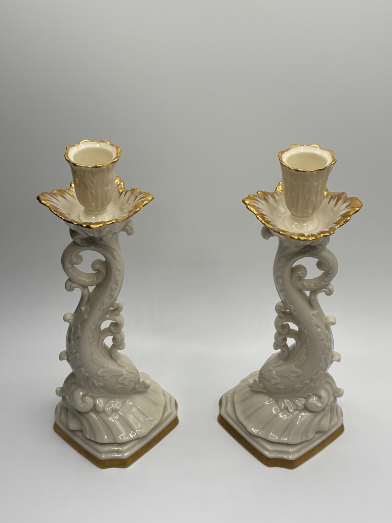 May include: A pair of ivory porcelain candle holders with gold accents. Each holder features a stylized dolphin design with a scalloped base.