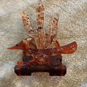 May include: A clear resin sculpture of a roadrunner and a saguaro cactus. The roadrunner is orange with glitter, and the cactus is filled with small stones. The sculpture is mounted on a rectangular base.