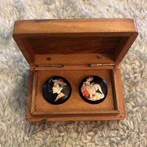 May include: A wooden box with an open lid, displaying two black circular pins. One pin shows a side profile of a person smoking, the other depicts a seated figure. The box is made of light brown wood.