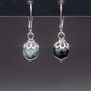 May include: A pair of dangle earrings with silver-tone metalwork. Each earring features a round, green and black speckled stone, suspended from a decorative silver-tone cap and a lever-back closure. The earrings are displayed against a dark background.