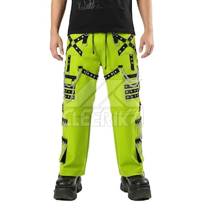 Może przedstawiać: Jasne neonowe zielone spodnie z czarnymi paskami, łańcuchami i detalami z przelotkami. Spodnie mają prosty krój nogawek i wiele kieszeni. Widoczny jest napis "LEERIK". Osoba ma na sobie czarne buty na platformie.