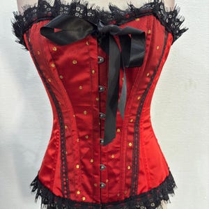 Puede incluir: Un corsé de satén rojo con ribete de encaje negro y un lazo de satén negro. El corsé tiene lunares dorados y un cierre de busk en la parte delantera.