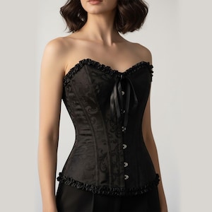 May include: Black corset with a floral design. The strapless top has a frilled trim, and a satin ribbon tie. The corset has metal clasps down the front. The garment is designed to cinch the waist.