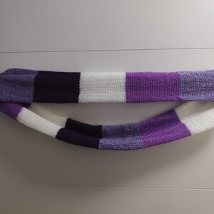 Könnte beinhalten: Ein gestrickter Loop-Schal mit einem Colorblock-Muster. Der Schal zeigt abwechselnd Blöcke in Weiß, verschiedenen Lilatönen und Schwarz. Der Schal ist aus einem weichen, strukturierten Garn gefertigt.