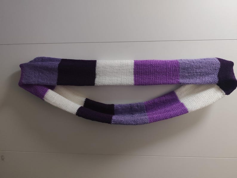 May include: A knitted infinity scarf in a color block pattern. The scarf features alternating blocks of white, various shades of purple, and black. The scarf is made of a soft, textured yarn.