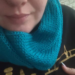 May include: A knitted turquoise cowl scarf. The scarf is made of a soft, textured yarn and is wrapped around the neck. The cowl is a vibrant shade of turquoise, adding a pop of color. The background is a black garment with a gold design.