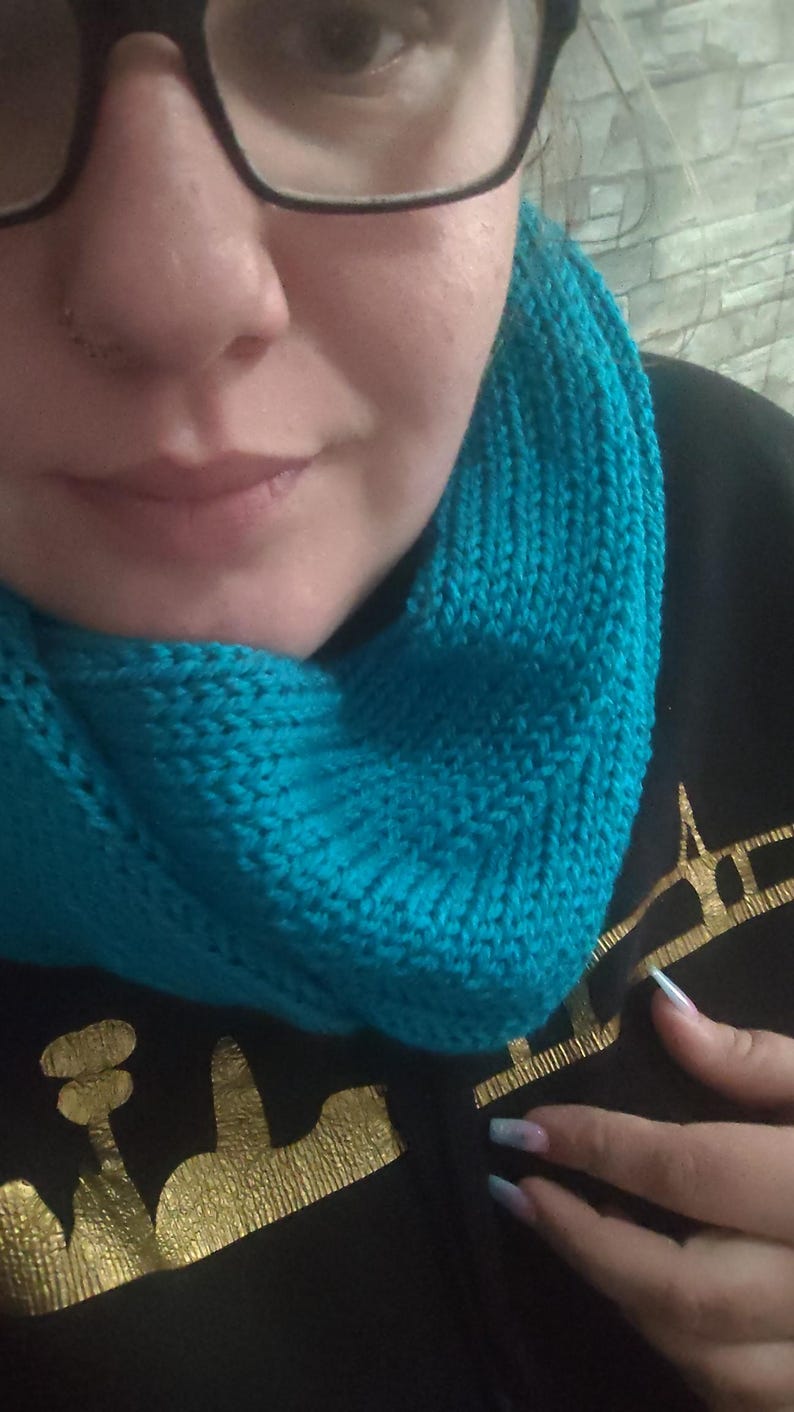 May include: A knitted turquoise cowl scarf. The scarf is made of a soft, textured yarn and is wrapped around the neck. The cowl is a vibrant shade of turquoise, adding a pop of color. The background is a black garment with a gold design.
