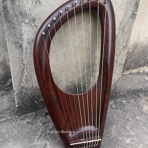 May include: A dark brown lyre harp with a curved frame and ten strings. The instrument has a polished finish and silver tuning pegs. The strings are a light gold color. The harp is set against a textured gray background. The text "Saliham Boutique" is visible.