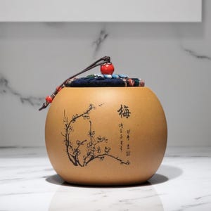 May include: A round, tan ceramic tea caddy with a black and white floral design and the Chinese character for "sea" on the front. The lid is covered in a blue and red fabric with a red tassel.