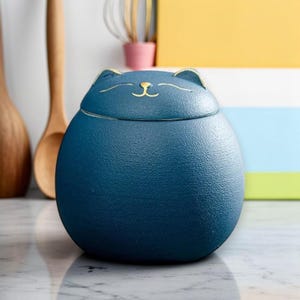 Könnte beinhalten: Ein dunkelblaues Keramikgefäß mit einem Katzengesicht-Design auf dem Deckel. Das Gefäß hat eine abgerundete Form und eine strukturierte Oberfläche. Das Katzengesicht ist mit goldenen Akzenten versehen. Das Gefäß wird wahrscheinlich zur Aufbewahrung oder Dekoration verwendet.