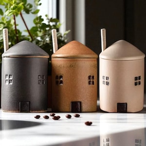 Peut inclure: Trois récipients en céramique en forme de maison, noirs, bruns et beiges. Chacun a un toit conique, des fenêtres carrées et une petite porte. Une cuillère blanche repose sur chaque couvercle. Des grains de café sont éparpillés devant.