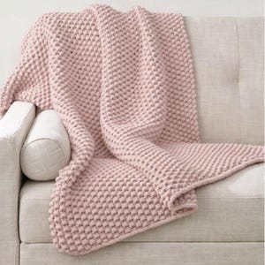 May include: A soft, pink knitted throw blanket draped over a light-colored sofa. The blanket has a textured, bobble-like pattern. The sofa has a neutral color and a button-tufted back. The blanket is folded over the arm of the sofa.