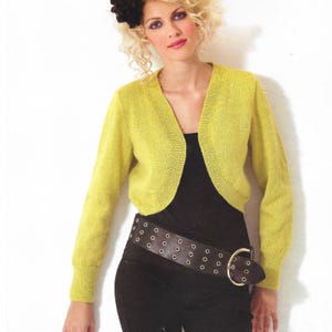 Puede incluir: Un bolero amarillo con mangas largas. La prenda se usa sobre un top negro y se combina con un cinturón negro ancho con ojales plateados y una hebilla dorada. La modelo lleva un accesorio para el cabello con forma de flor negra.