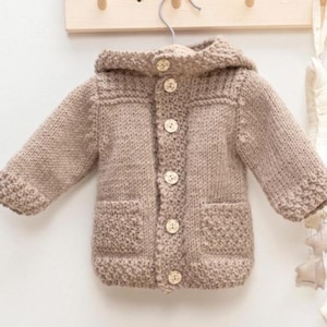 May include: A tan knitted baby cardigan with a hood, two front pockets, and wooden buttons. The cardigan is hanging on a metal hanger against a light-colored wall.
