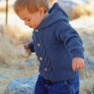 Può includere: Un cardigan blu con cappuccio lavorato a maglia con bottoni in legno. Il motivo a trecce corre lungo la parte anteriore del maglione. Il bambino indossa jeans blu.