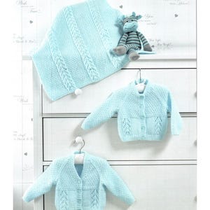 May include: A set of baby clothes in light blue, including a knitted blanket with cable detailing and two cardigans. A stuffed monkey with grey and blue stripes sits on the dresser. The cardigans have button fastenings.