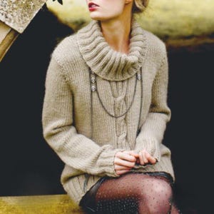 Può includere: Un maglione beige a trecce con collo alto ampio e a coste. Il maglione presenta un motivo a trecce sul davanti ed è abbinato a una lunga collana argentata. Lo stile generale è accogliente e caldo, adatto ai climi più freschi.