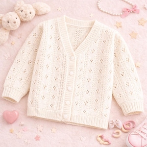 May include: Cream-colored, button-down cardigan sweater with a V-neck. The sweater has a delicate, open-knit pattern and ribbed cuffs and hem. The sweater is made of a soft, textured yarn. The background includes a pink surface and decorative items.
