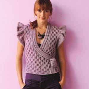 May include: A lavender-colored, knitted wrap top with short sleeves and a textured pattern. The top features a V-neck design and a tie closure at the waist. The sleeves have a ruffled detail. A necklace with dark beads is also visible.