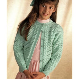 May include: A light green, open-weave cardigan with a cable knit design. The button-up sweater has a round neckline and long sleeves. The cardigan is worn over a pink dress. The girl is wearing a green bow in her hair.