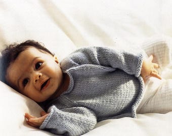 May include: A baby wearing a light blue knitted sweater and white pants, lying on a white surface. The sweater has long sleeves and a rounded neckline. The baby is looking at the camera.