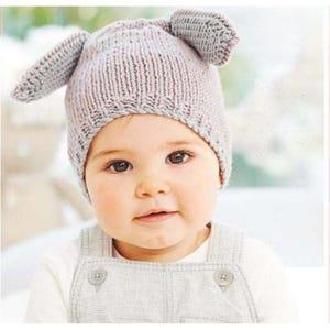 Puede incluir: Un bebé que lleva un gorro de punto gris claro con dos protuberancias en forma de orejas. El bebé también lleva una camiseta blanca de manga larga y un mono gris. El gorro y el mono están hechos de un material suave y texturizado.