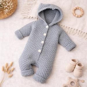 Könnte beinhalten: Hellblauer gestrickter Baby-Overall mit Kapuze und weißen Knöpfen. Der Overall hat lange Ärmel und Beine mit umgeschlagenen Bündchen. Ein Paar cremefarbene gestrickte Babyschuhe und ein Holzspielzeug sind ebenfalls zu sehen.