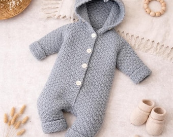 May include: A light blue knitted baby jumpsuit with a hood and white buttons. The jumpsuit has long sleeves and legs with folded cuffs. A pair of cream-colored knitted baby booties and a wooden toy are also visible.