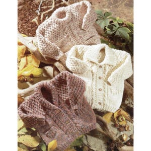 May include: Three hand-knitted sweaters in cream, tan, and mauve. The sweaters have button closures and varying textures, including cable knit and a textured stitch. The garments are displayed on a bed of autumn leaves and branches.