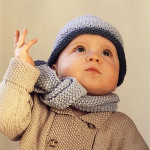 Puede incluir: Un bebé con gorro, bufanda y cárdigan de punto. El gorro es gris claro con un borde azul oscuro. La bufanda y el cárdigan son gris claro y beige, respectivamente. El bebé mira hacia arriba con la mano levantada.