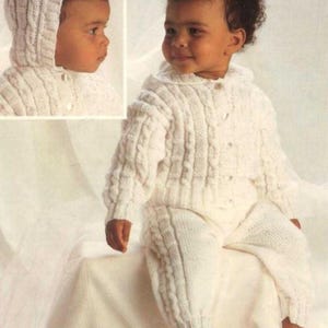 May include: A knitted white baby outfit, featuring a hooded jacket and matching pants. The outfit has a cable knit design and button closures. The baby is wearing the outfit. The outfit is made of a soft, warm material, perfect for keeping a baby cozy.